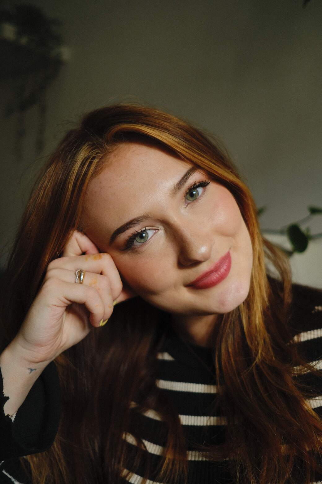 A portrait of a young woman with long, brown hair and green eyes she wears a black top with white stripes and has her hand resting on her head Her lips are red and glossy The background is blurred, revealing an indoor wall with a textured pattern She wears a glinting ring on her left hand
