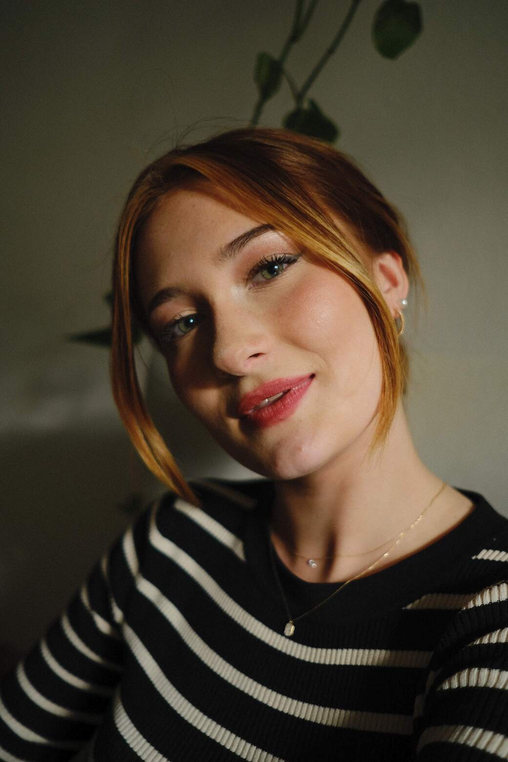 A young woman with brown hair that is tied into a ponytail wearing a black shirt with white stripes Her smile reveals white teeth and her head is slightly tilted The background is blurred, revealing an indoor wall with a textured pattern also you can see a delicate necklace that shimmers against her pale skin